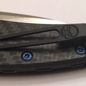 Op de afbeelding: Een zwarte zakclip met een schedelontwerp bevestigd aan een zwart en grijs carbonfiber meshendel. De clip heeft een blauwe schroef aan de rechterkant en een blauwe schroef aan de linkerkant.