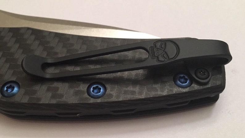 Puede incluir: Un clip de bolsillo negro con un dise&ntilde;o de calavera unido a un mango de cuchillo de fibra de carbono negro y gris. El clip tiene un tornillo azul en el lado derecho y un tornillo azul en el lado izquierdo.