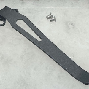 May include: Black metal pocket clip with a rounded end and a small hole on the side. The clip is designed to attach to a pocket or belt.