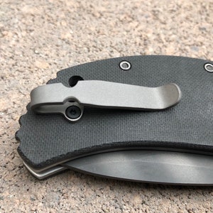 May include: A black pocket knife with a titanium clip and a textured handle. The knife has a partially open blade.