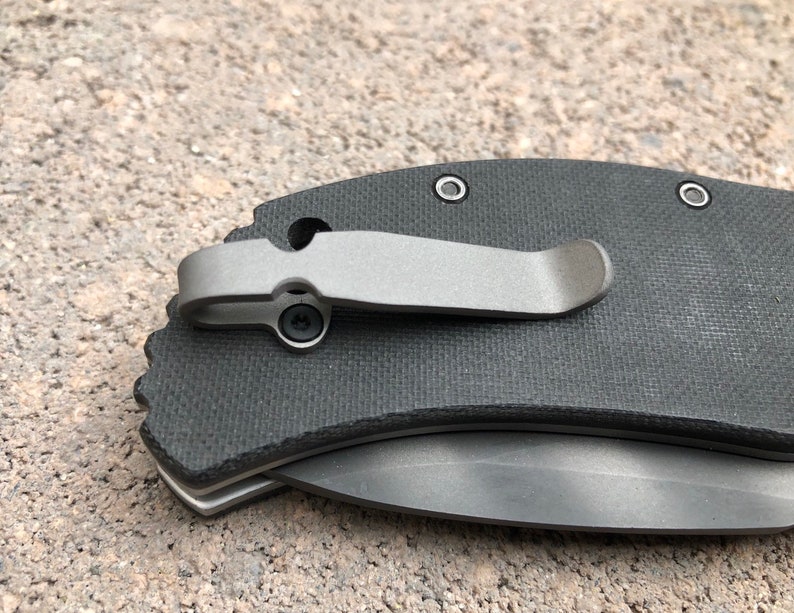 May include: A black pocket knife with a titanium clip and a textured handle. The knife has a partially open blade.