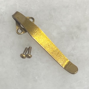 May include: A gold-toned metal pocket clip with a curved design and a triangular mounting base featuring two circular screw holes. The clip is accompanied by two small silver-colored metal screws with threaded shanks and rounded heads.