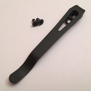 May include: Black metal pocket clip with two screws. The clip is curved and has a hole in the top.