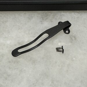 May include: Black metal pocket clip with two screws. The clip is curved and has a small hole in the center.