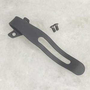 May include: A matte black pocket clip with two small screws. The clip has a curved design with a long oval cutout and two mounting holes. The screws are silver and have a slotted head.