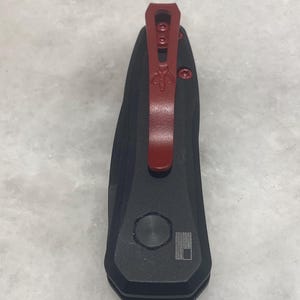 May include: A black pocket knife with a red pocket clip. The knife has a small, round button on the side and a small, engraved American flag.