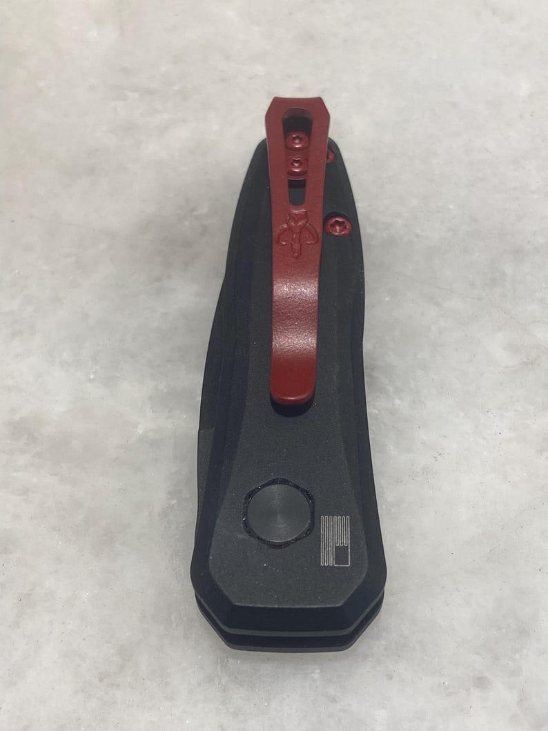 May include: A black pocket knife with a red pocket clip. The knife has a small, round button on the side and a small, engraved American flag.