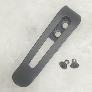 Può includere: Una clip da tasca in metallo nero opaco con un lungo intaglio stretto e due fori di montaggio circolari a un'estremità. La clip è accompagnata da due piccole viti nere a testa piatta, ciascuna dotata di un'impronta a stella per il fissaggio.