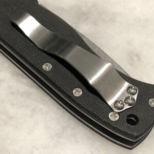 May include: Close-up of a black pocket knife with a stainless steel clip. The clip is attached to the knife with four screws.