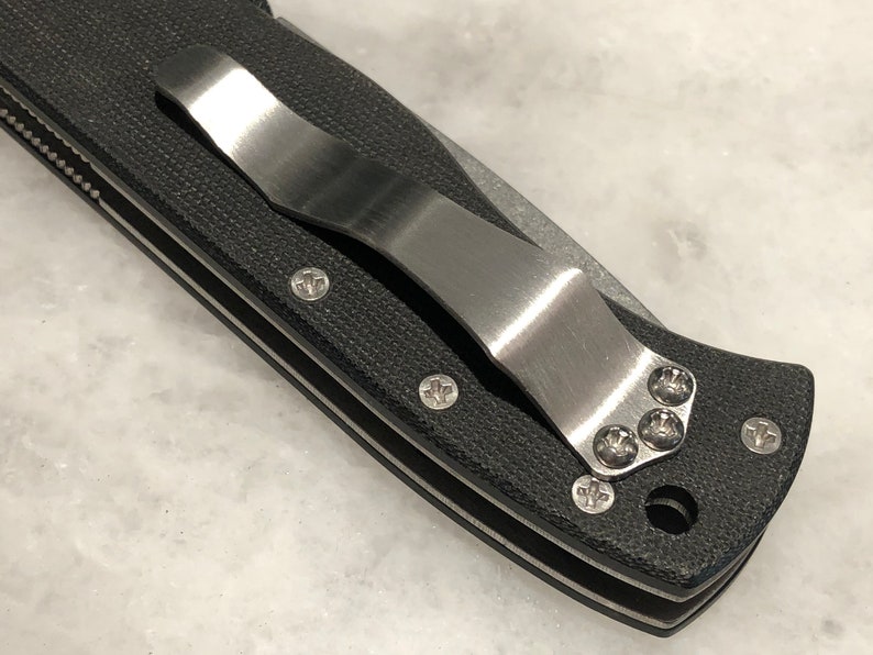 May include: Close-up of a black pocket knife with a stainless steel clip. The clip is attached to the knife with four screws.