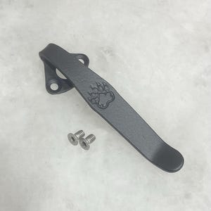 May include: A matte black metal pocket clip featuring an embossed bear paw print design. The clip has a triangular mounting base with two circular screw holes and is accompanied by two small silver-colored metal screws with flat heads.