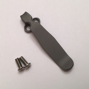 May include: A grey metal pocket clip with two screws. The clip has a curved shape and a smooth finish.