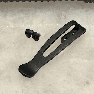 May include: Black metal pocket clip with two small black screws. The clip has a rounded end and a rectangular opening.