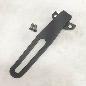 May include: A black metal clip with a curved top and a long, slender body. Two small screws are included. The clip has a hole in the center and two holes at the top. The clip is designed for securing items.
