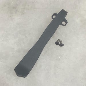 May include: A dark gray pocket knife clip with two screw fasteners. The clip is a long, slender piece with a curved design and two holes at one end for attachment. The screws are small and black.