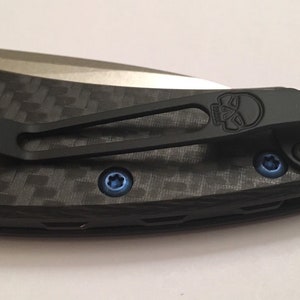 Puede incluir: Un clip de bolsillo negro con un dise&ntilde;o de calavera unido a un mango de cuchillo de fibra de carbono negro y gris. El clip tiene tornillos azules.