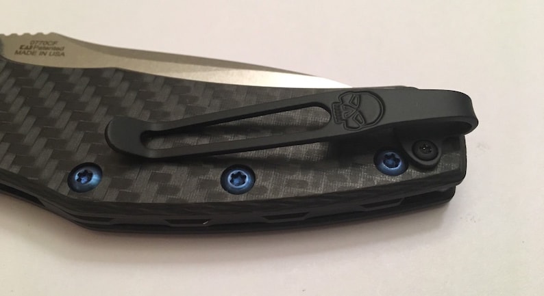 Puede incluir: Un clip de bolsillo negro con un dise&ntilde;o de calavera unido a un mango de cuchillo de fibra de carbono negro y gris. El clip tiene tornillos azules.