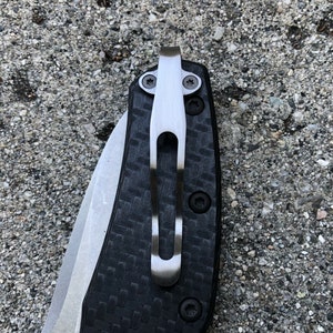 May include: A close-up of a black and silver pocket knife with a carbon fiber handle and a deep carry clip.