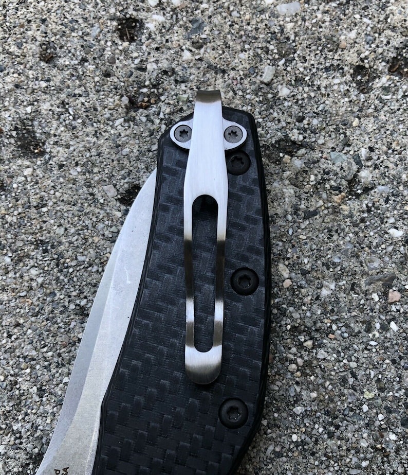 May include: A close-up of a black and silver pocket knife with a carbon fiber handle and a deep carry clip.