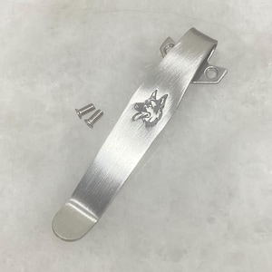 May include: A brushed silver metal pocket clip featuring an engraved German Shepherd dog head profile. The clip has a curved design with a mounting base containing two screw holes, accompanied by two small matching silver metal screws for attachment.