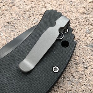 May include: A close-up of a black pocket knife with a silver pocket clip. The clip is attached to the knife with two screws.