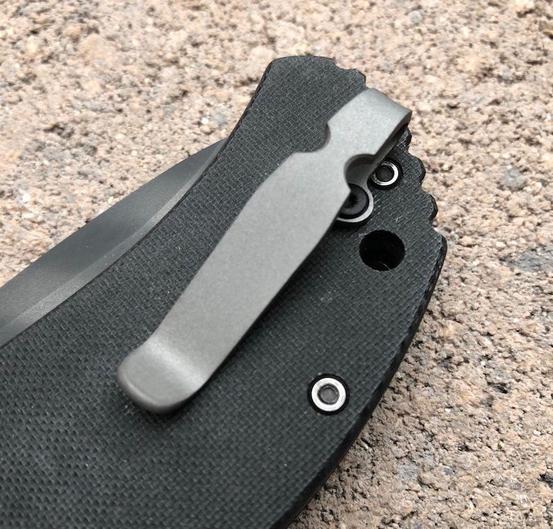 May include: A close-up of a black pocket knife with a silver pocket clip. The clip is attached to the knife with two screws.