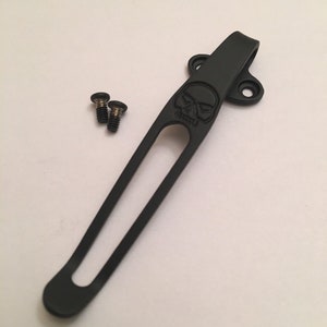 Puede incluir: Clip de bolsillo de metal negro con un dise&ntilde;o de calavera y dos tornillos.