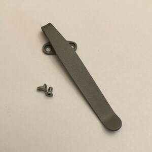 May include: A gray titanium pocket clip with two screws. The clip is designed to attach to a pocket knife or other tool.