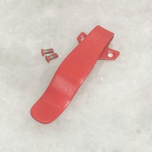 May include: A bright red metal clip with two small red screws. The clip has a curved design with two holes on one end for mounting. The screws are small and have a slotted head. The items are on a light gray surface.