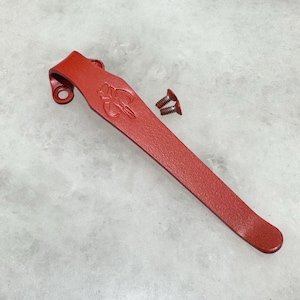 Puede incluir: Un clip de bolsillo de metal rojo con un acabado texturizado. El clip tiene un pequeño logotipo en relieve. Se incluyen dos tornillos pequeños.