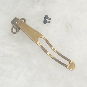 May include: A brass-colored pocket knife clip with two small black screws. The clip has a curved, elongated design with a hole at the top for attachment and a slotted section for securing the knife. The metal has a slightly worn appearance.