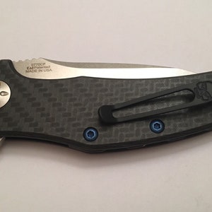 Puede incluir: Un cuchillo de bolsillo negro y plateado con un mango de fibra de carbono y un clip negro. El cuchillo tiene un logotipo de calavera en el clip y el texto "0770CF KAI Patented MADE IN USA" en el mango.
