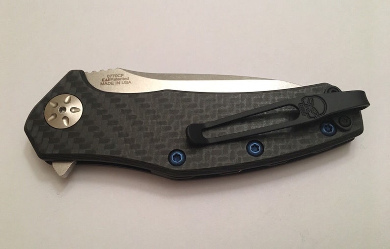 Puede incluir: Un cuchillo de bolsillo negro y plateado con un mango de fibra de carbono y un clip negro. El cuchillo tiene un logotipo de calavera en el clip y el texto "0770CF KAI Patented MADE IN USA" en el mango.