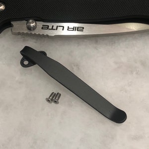 May include: Black metal pocket clip with two silver screws. The clip is designed to attach to a pocket knife. The text "Air Lite" is visible on the knife blade.