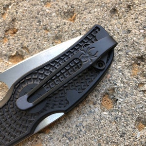 Könnte beinhalten: Nahaufnahme eines schwarzen Taschenmessers mit einem strukturierten Griff und einem Spinnenlogo am Clip.