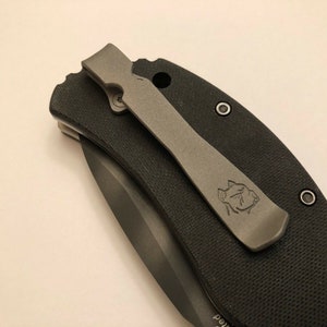 Puede incluir: Un cuchillo de bolsillo negro con un clip de bolsillo gris. El clip de bolsillo tiene un peque&ntilde;o logotipo.