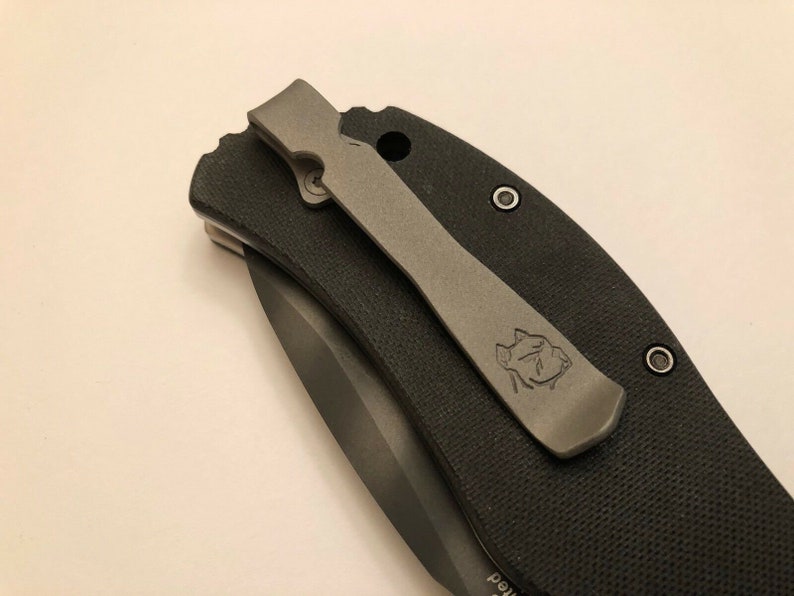 Puede incluir: Un cuchillo de bolsillo negro con un clip de bolsillo gris. El clip de bolsillo tiene un peque&ntilde;o logotipo.