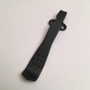 May include: A black metal pocket clip with a three-leaf clover design. The clip has two holes for attaching to a belt or pocket.