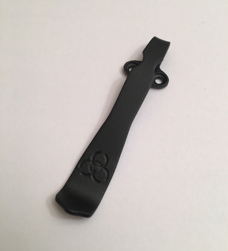 May include: A black metal pocket clip with a three-leaf clover design. The clip has two holes for attaching to a belt or pocket.
