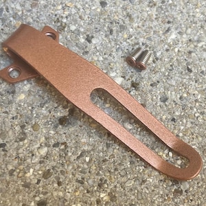 May include: A copper-colored metal pocket clip with a long, narrow cutout and two mounting holes at the top, accompanied by two small silver-toned metal screws. The clip has a smooth, matte finish and a curved design for attachment.