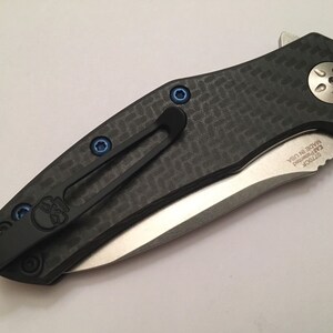 Puede incluir: Un cuchillo de bolsillo negro y plateado con un mango de fibra de carbono y un tornillo azul. El cuchillo tiene un clip y el texto "ZT 0770CF" y "MADE IN USA" est&aacute;n grabados en la hoja.