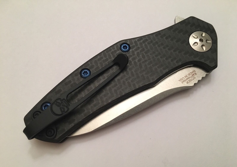 Puede incluir: Un cuchillo de bolsillo negro y plateado con un mango de fibra de carbono y un tornillo azul. El cuchillo tiene un clip y el texto "ZT 0770CF" y "MADE IN USA" est&aacute;n grabados en la hoja.