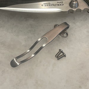 May include: A silver-colored pocket knife clip with two small screws. The clip has a curved design with an open center. The knife blade is visible in the background.