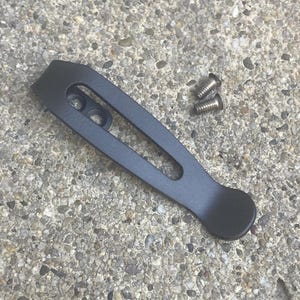 May include: A matte black metal pocket clip with a long, narrow cutout and two circular mounting holes at the top, accompanied by two small silver-colored metal screws with threaded shanks, designed for attachment to a tool or accessory.