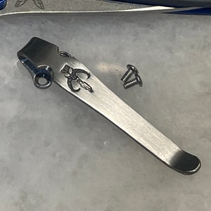 May include: A silver pocket clip with a small, engraved design. Two silver screws are included.