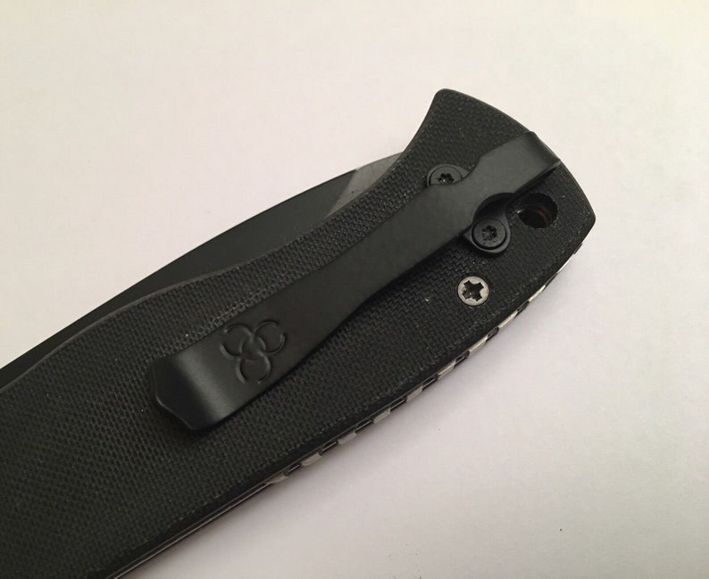 May include: Black pocket knife with a clip and a textured handle. The clip has a logo of three overlapping circles.