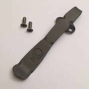 May include: A gray metal pocket clip with a skull design. The clip has two holes for attaching screws. Two screws are included.