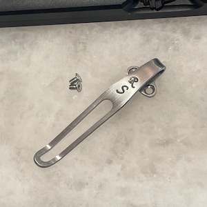 May include: A silver pocket clip with a rounded end and a small loop at the top. The clip is engraved with the letters "R.S.". Three small silver screws are also included.