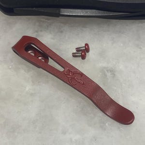 May include: A red metal pocket clip for a knife. The clip has a curved shape and a small hole on the top. Two small screws with red heads are included.