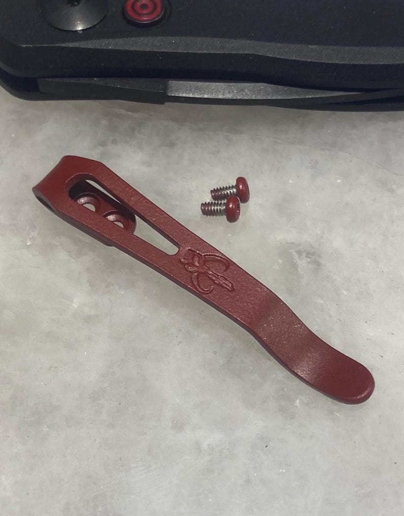 May include: A red metal pocket clip for a knife. The clip has a curved shape and a small hole on the top. Two small screws with red heads are included.